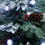Close-up of a Christmas tree branch decoration with green pine needles, white faux snow, brown pinecones, red berries, and glowing white string lights.