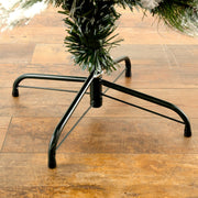 A close-up of a black metal Christmas tree stand supporting a green artificial tree, set on a brown wooden floor.