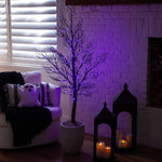 A bare decorative tree in a white planter, lit with purple string lights, placed in a cozy room beside a white armchair, pillows, and black lanterns with candles. A white brick fireplace wall is visible in the background.