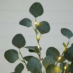 Eucalyptus branch string lights with glowing warm white fairy lights, showing realistic green leaves with detailed vein patterns against a neutral light gray background.