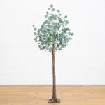 An artificial eucalyptus tree with a realistic brown trunk and round silver-green leaves, standing on a wooden floor against a white paneled wall.