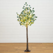 A tall artificial eucalyptus tree with glowing warm white LED fairy lights woven through the green leaves, standing on a wooden floor against a white shiplap wall.