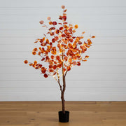An artificial autumn tree with orange and red leaves on a brown trunk, potted in a black plastic container, set against a white paneled wall and light wood floor.