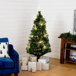 A decorated artificial Christmas tree with warm white lights stands in a corner of a room with white shiplap walls and a light wood floor. Several wrapped gift boxes are placed at the base of the tree, with a blue armchair on the left and a wooden side table on the right.
