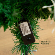 A close-up of a black plastic Christmas tree controller tag hanging among green artificial tree branches, with a wood surface visible in the background and the text '12 Functions Available' in the lower right.