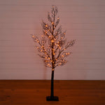 A bare artificial tree with a dark brown trunk and branches, decorated with warm glowing fairy lights, standing on a black base against a white shiplap wall and wooden floor.