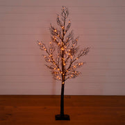 A bare artificial tree with a dark brown trunk and branches, decorated with warm glowing fairy lights, standing on a black base against a white shiplap wall and wooden floor.