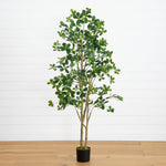 An artificial potted tree with green oval leaves and brown branches, standing in a black pot on a light wood floor against a white paneled wall.