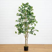 An artificial potted tree with green oval leaves and brown branches, standing in a black pot on a light wood floor against a white paneled wall.