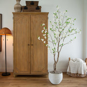 A tall wooden cabinet stands against a white wall in a styled room, next to a potted flowering tree with white blossoms in a white pot. A floor lamp is on the left and a woven chair with a cream throw blanket is on the right.