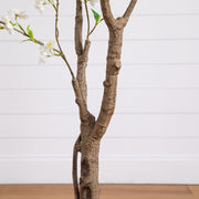 A decorative faux tree branch arrangement with textured brown bark, small green leaves, and white blossoms, shown against a white paneled wall and light wood floor.