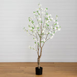 An artificial flowering tree with white blossoms and green leaves on a black pot, set against a white paneled wall and light wood floor.