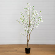 An artificial flowering tree with white blossoms and green leaves on a black pot, set against a white paneled wall and light wood floor.