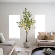 A tall potted artificial olive tree with green and yellow leaves stands in a bright, modern living room between two large windows, flanked by gray sofas with neutral-toned cushions.