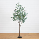 An artificial eucalyptus tree with green leaves and brown branches, standing on a black rectangular base against a white shiplap wall and light wood floor.