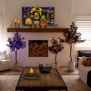 A cozy living room scene featuring a large framed Halloween-themed art print with colorful jack-o'-lantern houses and ghosts, hung on a white brick fireplace mantel above a wood fire. The room is decorated with three potted artificial trees, a wooden coffee table, and a white sofa.