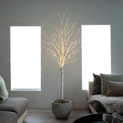 A decorative artificial tree with a white faux-birch trunk and delicate warm white string lights, displayed in a textured gray pot in a modern living room setting.