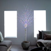 A decorative artificial tree with a white faux-birch trunk and many thin branches lit with multicolored LED fairy lights, placed in a gray woven pot in a modern living room setting.