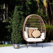 A woven rattan egg-shaped outdoor chair with white cushions and decorative throw pillows, set on a patio in front of a pond with lush green trees.