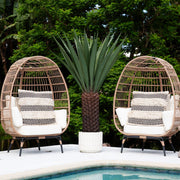 Two wicker chairs with white cushions and a potted plant on a poolside deck.
