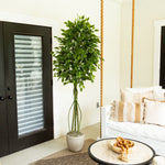 A tall artificial green tree in a light gray pot, styled in a bright, modern entryway next to a black door and a white daybed with rope accents.
