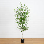 A tall artificial birch-style tree with green leaves and a white trunk, potted in a black cylindrical planter, standing on a light wood floor against a white paneled wall.