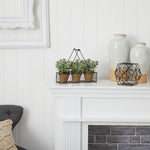 Decorative mantel with potted plants, vases, and a geometric candle holder against a white paneled wall.