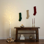 A styled holiday interior scene with a dark wooden console table against a beige wall, three knitted Christmas stockings hanging on the wall, a glowing artificial birch tree with logs at its base, candles, and decorative boxes on the table and shelf.
