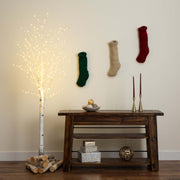 A styled holiday interior scene with a dark wooden console table against a beige wall, three knitted Christmas stockings hanging on the wall, a glowing artificial birch tree with logs at its base, candles, and decorative boxes on the table and shelf.
