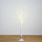 A lit artificial birch tree with white faux bark and warm white LED fairy lights along the branches, standing on a white base against a white paneled wall and light wood floor.