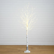 A lit artificial birch tree with white faux bark and warm white LED fairy lights along the branches, standing on a white base against a white paneled wall and light wood floor.