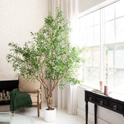 A tall artificial olive tree with green leaves and small yellow olives in a white pot, placed in a bright living room next to a window and a black console table.