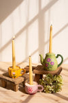 Three decorative taper candles in yellow, pink, and green floral ceramic holders, styled on small wooden blocks with a dried green flower sprig against a sunlit cream wall.