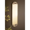 A tall, narrow rectangular wall mirror with a decorative metallic frame featuring leaf motifs at the top and bottom, mounted on a dark brown wall with some green foliage visible on the left.