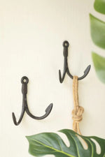Two black anchor-shaped wall hooks on a light cream wall, with a natural jute rope knotted and hung on the right hook. Green monstera leaves are partially visible in the foreground and background.