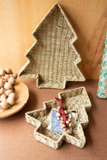 Two woven natural-fiber decorative trays shaped like a Christmas tree and a star, styled on a brown surface with a wooden bowl of wooden beads and a small gift box in the foreground.