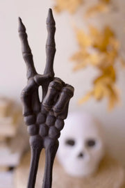 A dark gray decorative skeleton hand prop holding up a peace sign, set against a softly blurred background with a white skull and warm yellow dried leaves.
