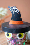 A Halloween-themed candy jar with a black lid topped by a black witch hat decoration with white spiderwebs and orange accents.