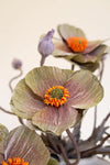 A close-up of artificial anemone-style flowers with muted olive-green and dusty rose petals, bright orange centers, and soft purple buds on a plain light background.
