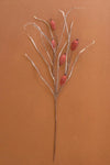 A decorative artificial branch with thin light beige twigs and several small textured reddish-brown berry-like accents, photographed against a solid warm brown background.