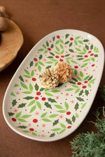 An oval white ceramic serving platter decorated with a green holly and red berry pattern, styled with two small pinecones on a brown surface.