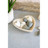 A hand carved wooden heart-shaped bowl filled with smooth stones on a grey surface, with a woven mat and a palm leaf in the background.